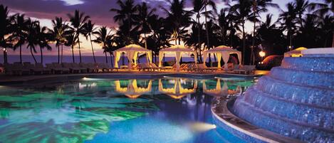 Ein Pool mit klarem, blauem Wasser, umgeben von Liegestühlen und Palmen, mit einem Sonnenuntergang im Hintergrund.