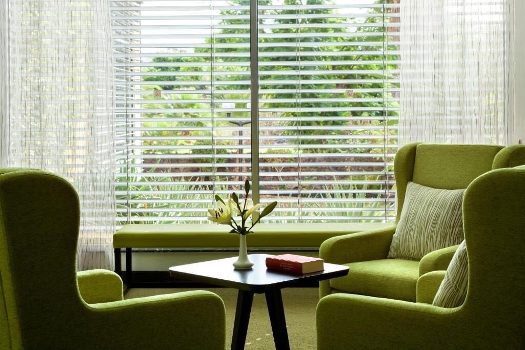 Ein Zimmer mit einem grünen Sofa, einem kleinen Tisch mit einer Vase und einem Buch sowie einem Fenster mit Jalousien.