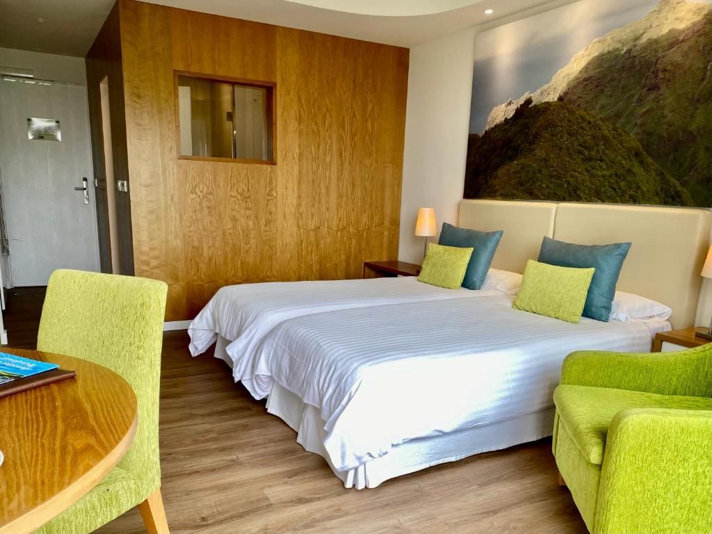 A hotel room with a bed, two nightstands, a lamp, a chair, a table, and a painting on the wall.