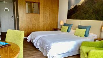 A hotel room with a bed, two nightstands, a lamp, a chair, a table, and a painting on the wall.