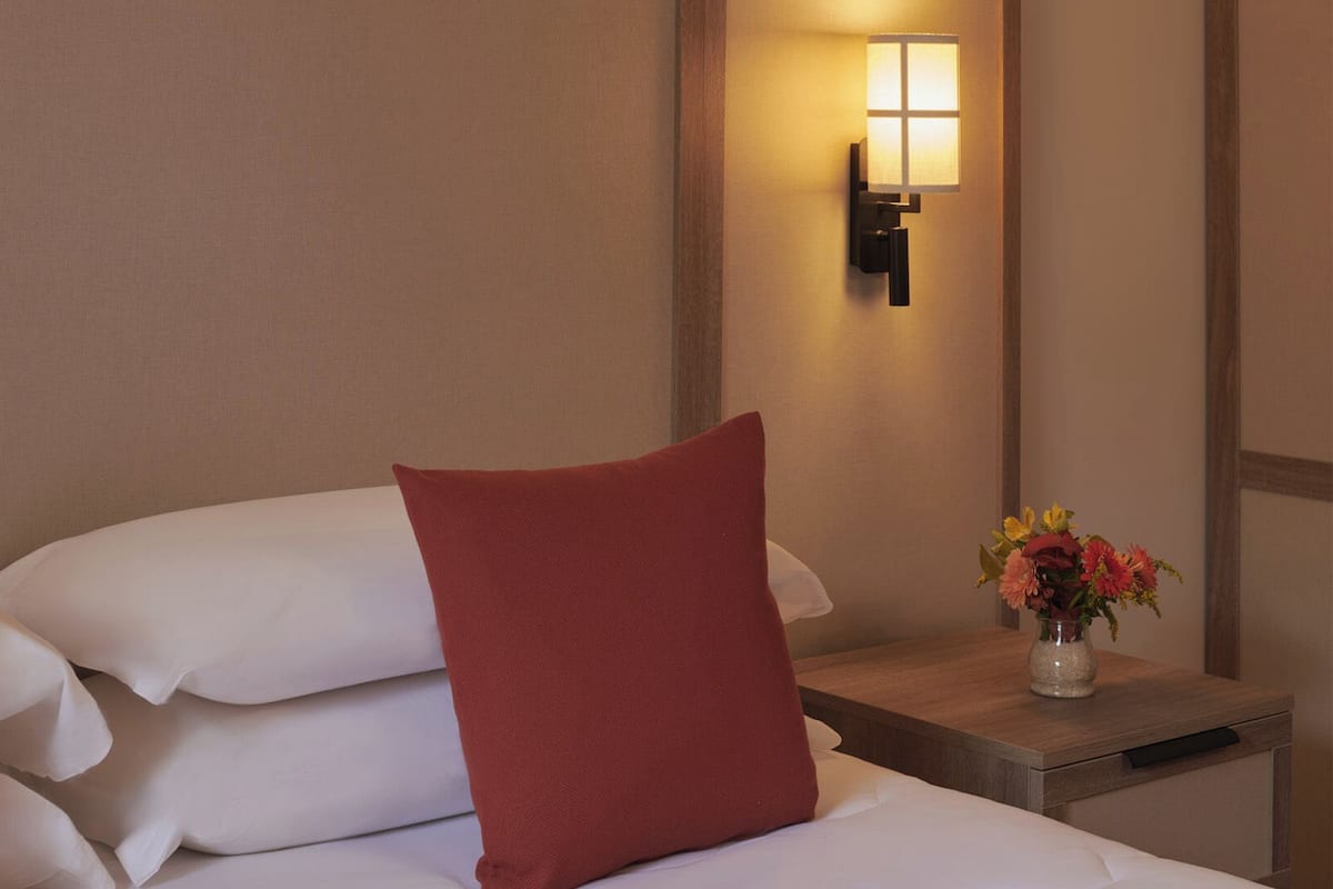 Ein Hotelzimmer mit Bett, Kissen, einem Nachttisch, einer Vase mit Blumen und einer Wandlampe.