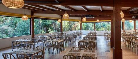 A spacious dining area with wooden tables and chairs, large windows offering a view of greenery, and hanging pendant lights.