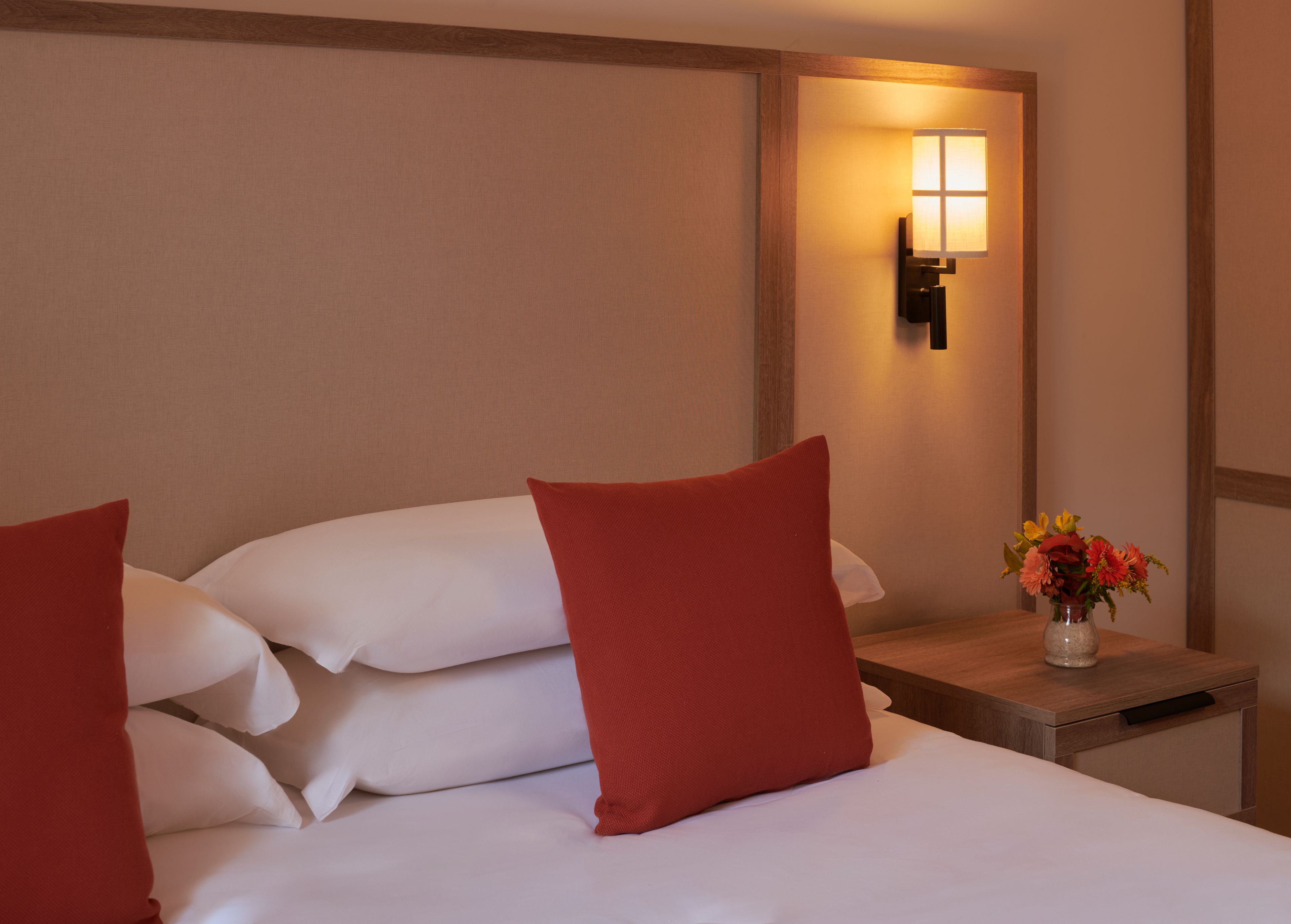 Una habitación de hotel con una cama bien hecha, una mesita de noche con un florero y una lámpara de pared.