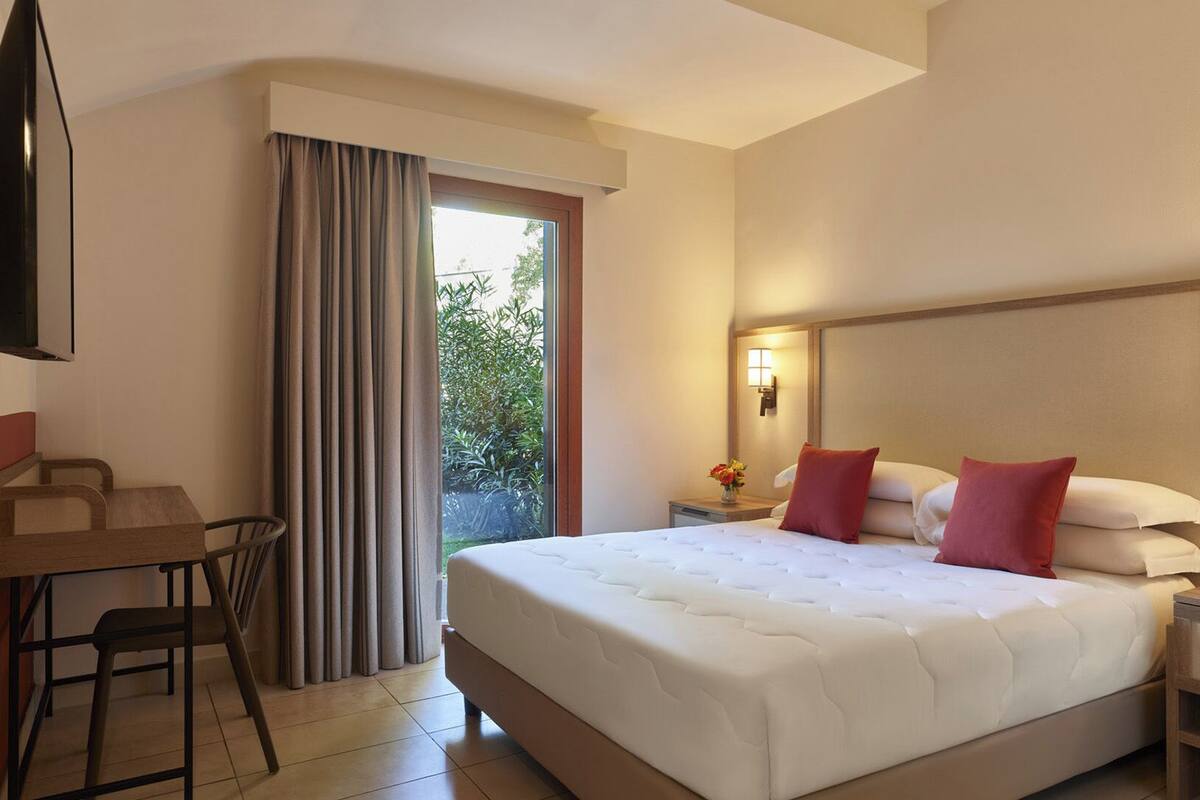 Ein Hotelzimmer mit einem großen Bett, einem Schreibtisch mit Stuhl, einem an der Wand befestigten Fernseher und einem Fenster mit Vorhängen.