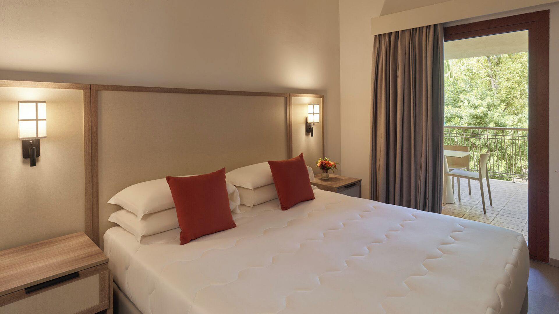 Una camera d'albergo con un letto grande, due cuscini rossi, un comodino con una lampada e vista su un balcone con mobili da esterno.