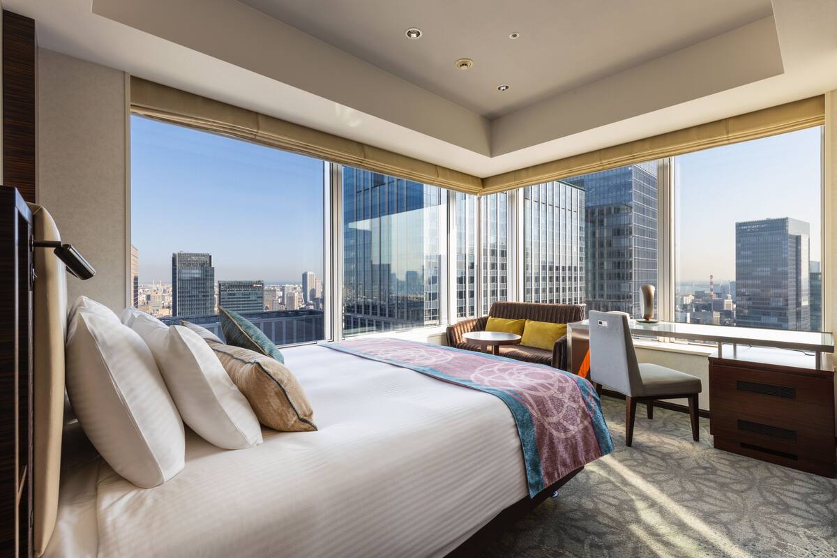 A hotel room with a large bed, a desk, a chair, and a view of the cityscape through large windows.