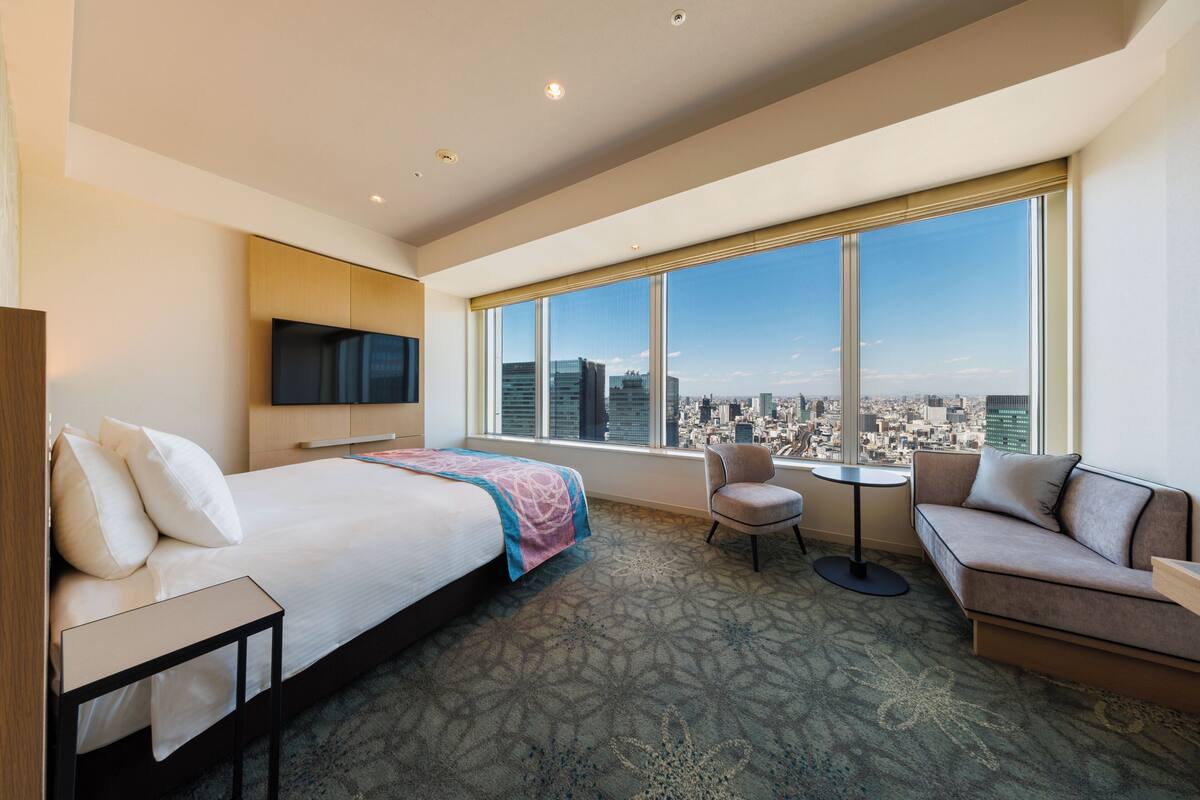 A hotel room with a bed, a sofa, a chair, a small table, and a TV mounted on the wall.