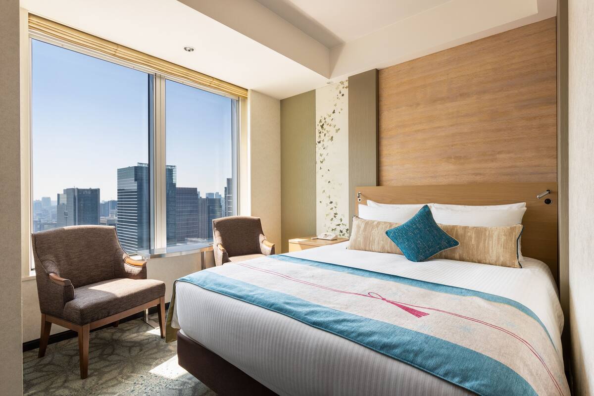 A hotel room with a large bed, a chair, a nightstand, and a view of the city skyline through a large window.