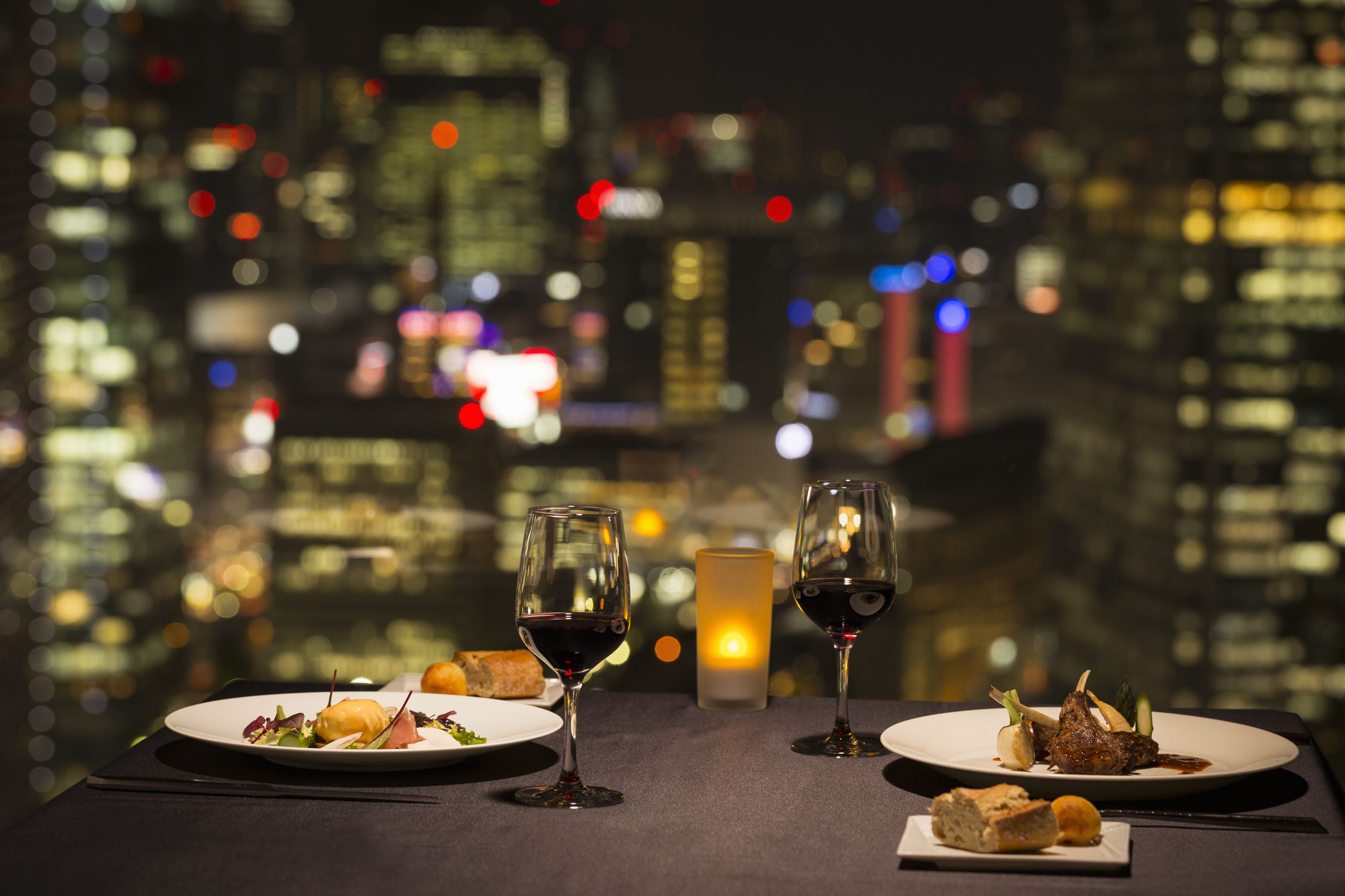Mesa lista para cenar con dos copas de vino, una vela y platos de comida, todo sobre un fondo de la ciudad.
