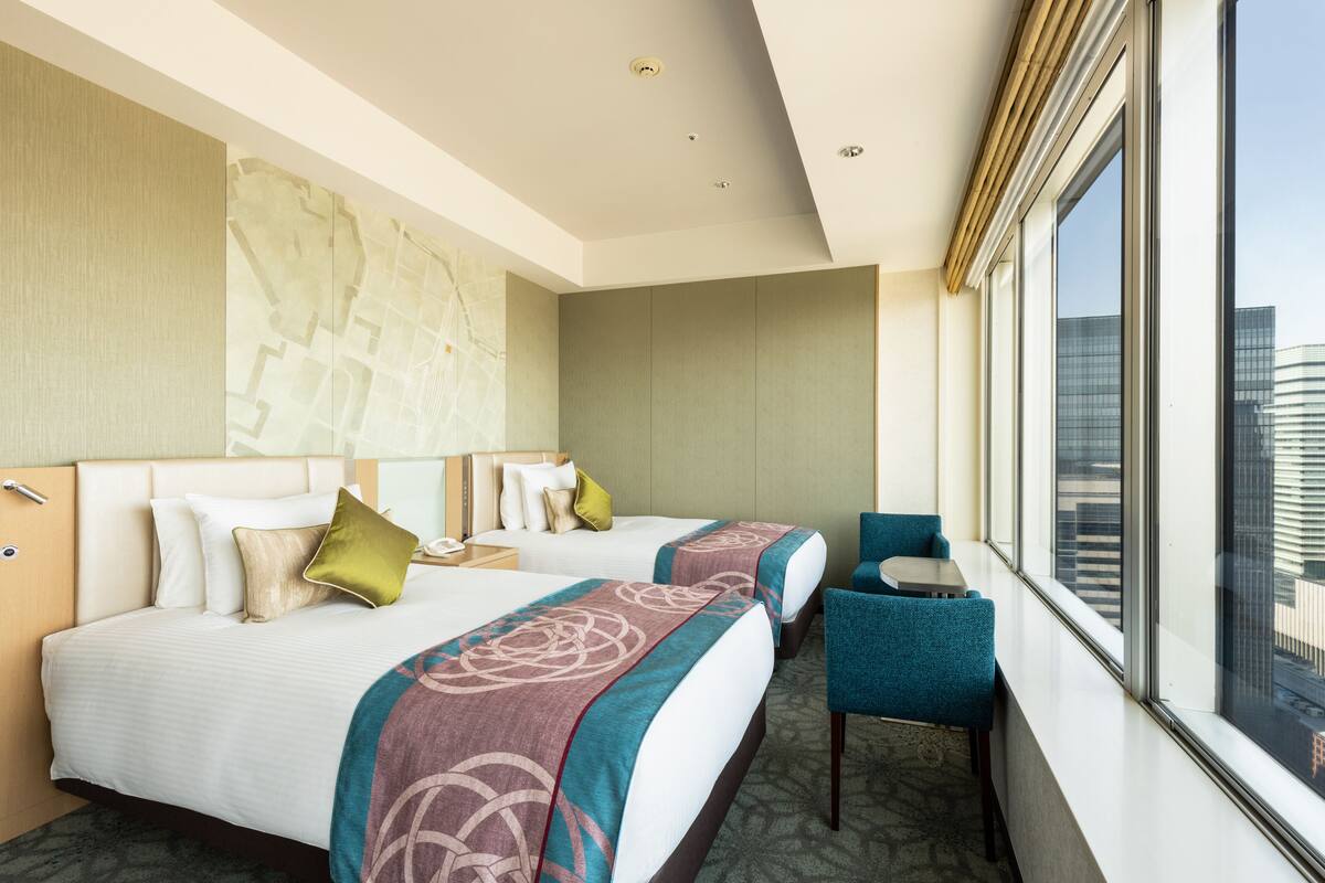 A hotel room with two beds, a small table, a chair, and a large window offering a city view.