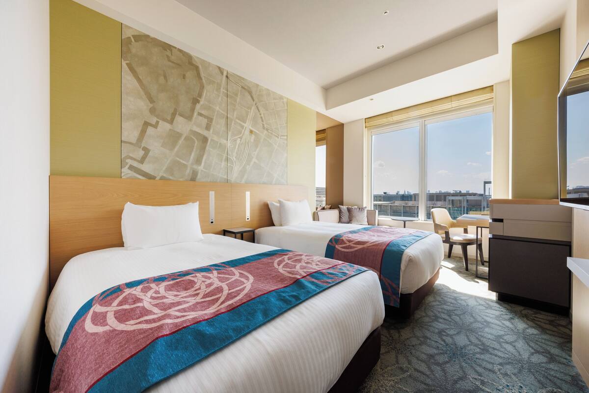 A hotel room with two beds, a desk, and a large window offering a city view.