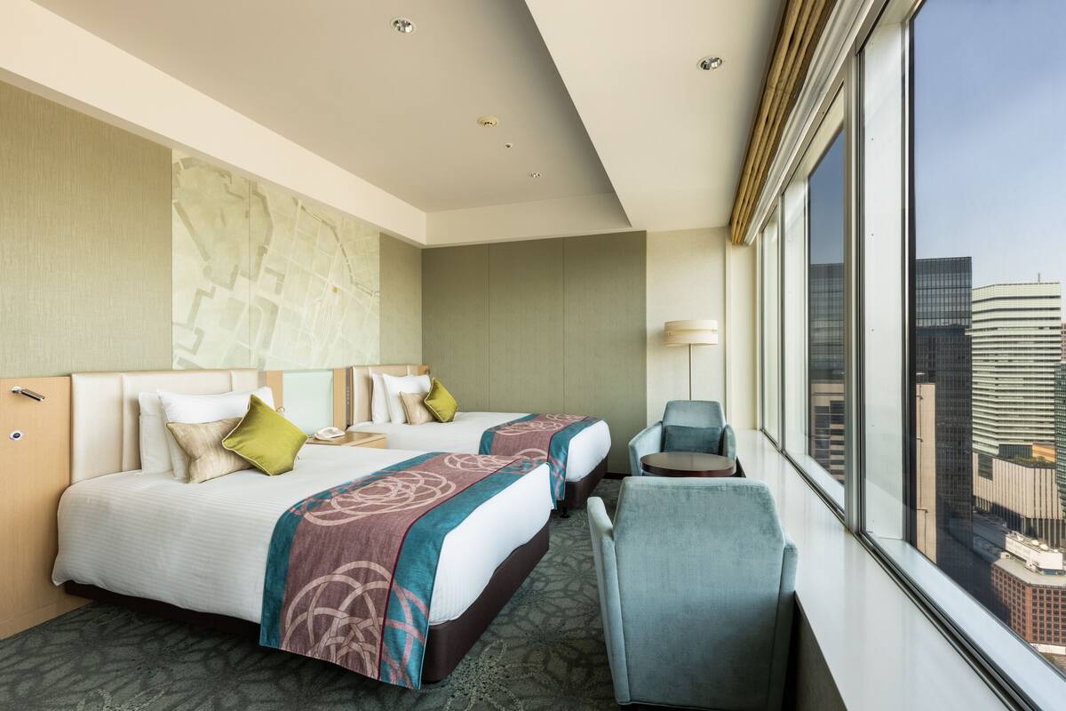 A hotel room with two beds, a desk, a chair, and a large window offering a city view.