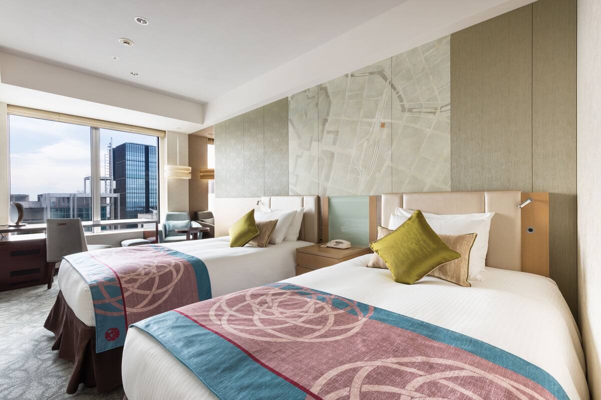 A hotel room with two beds, a desk, and a large window with a city view.