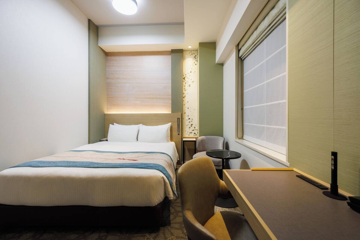 A hotel room with a bed, a desk, a chair, a small table, and a window.