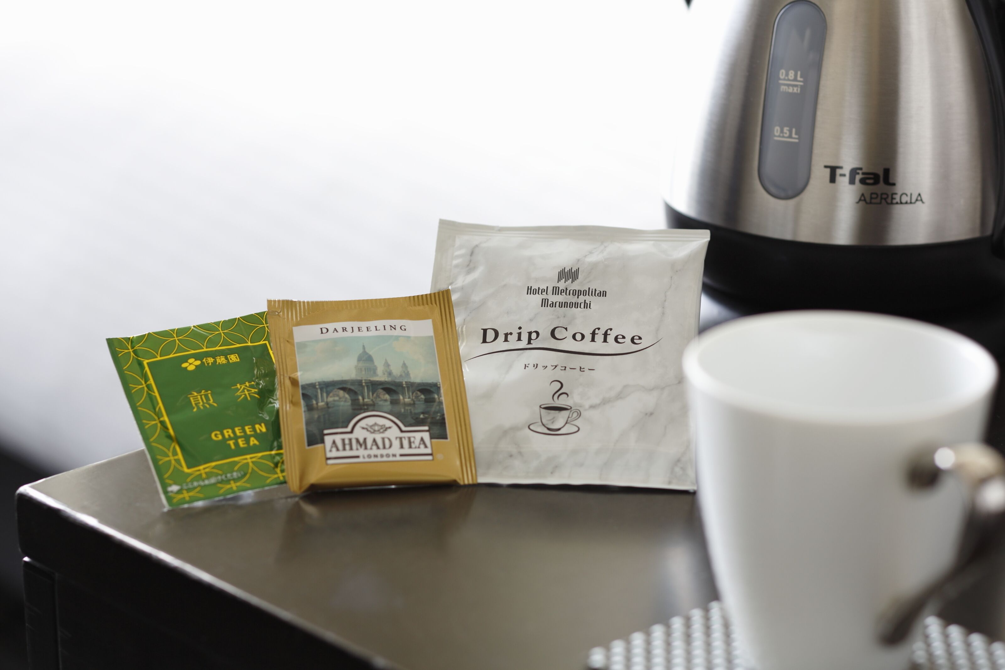 A coffee maker, a drip coffee packet, a green tea packet, and a white mug on a table.