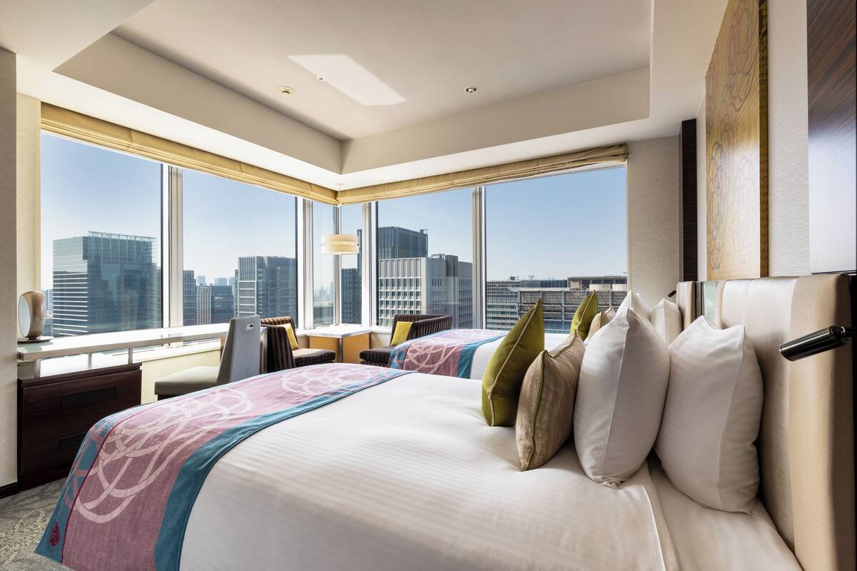 A hotel room with a large bed, two bedside tables, a desk, and a chair. There are large windows offering a city view.