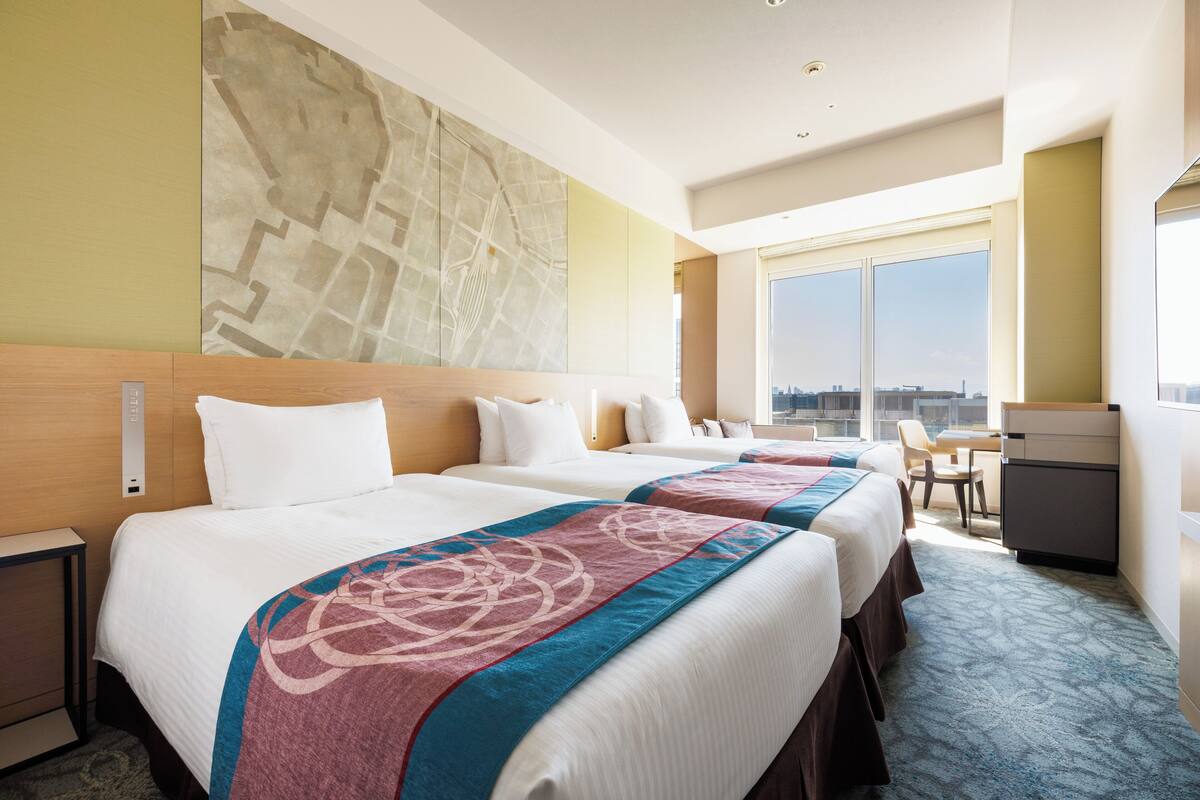A hotel room with two beds, a desk, a chair, and a large window offering a city view.