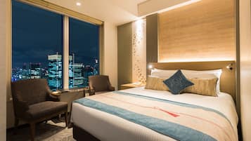 A hotel room with a large bed, a chair, a nightstand, and a view of the city at night.