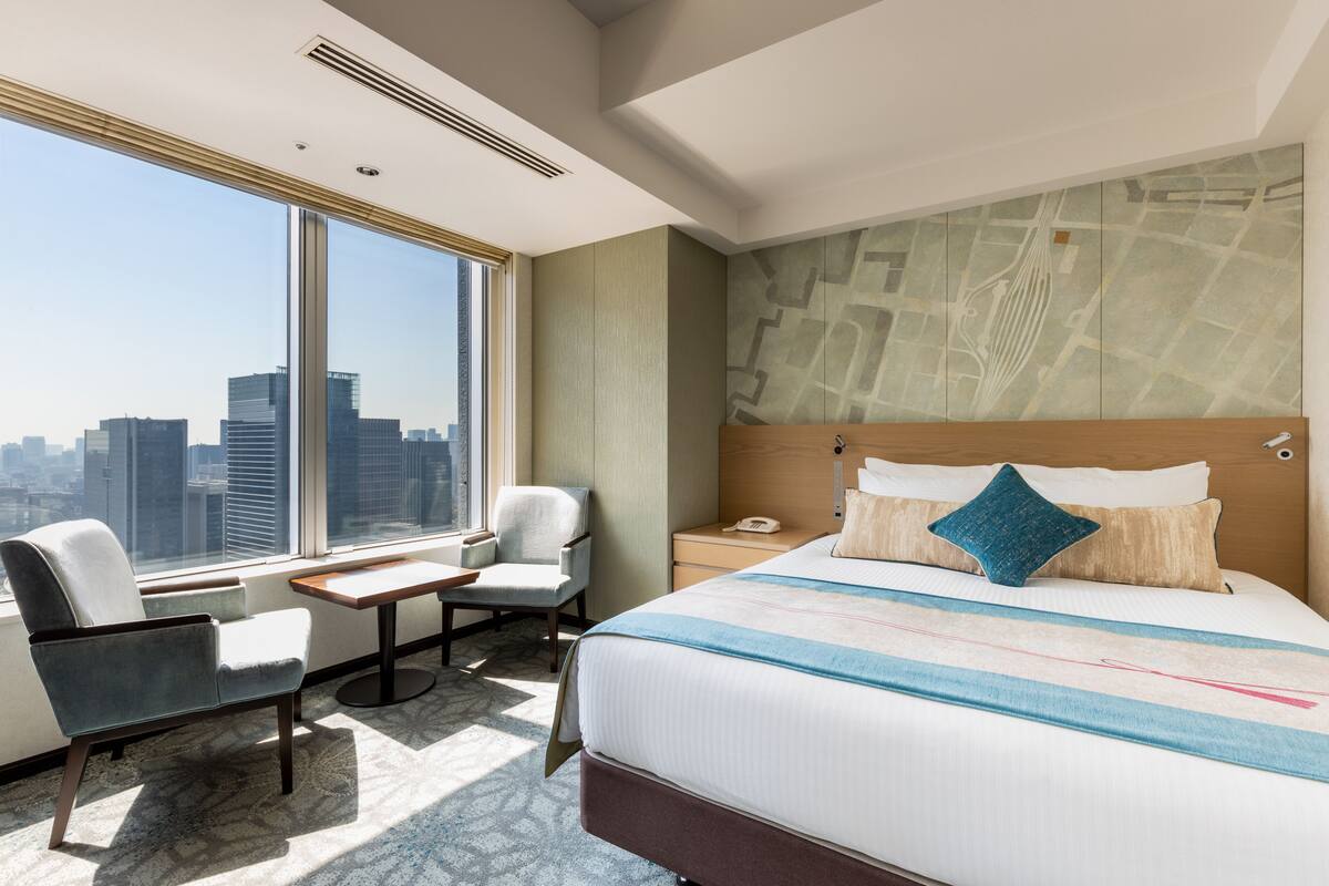 A hotel room with a large bed, a desk, two chairs, a nightstand, and a city view.