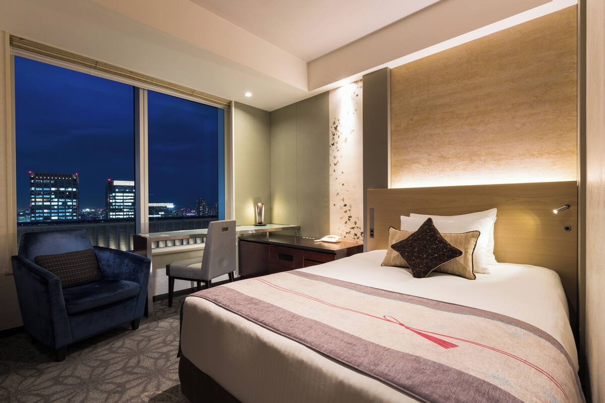 A hotel room with a large bed, a desk, a chair, and a city view through the window.