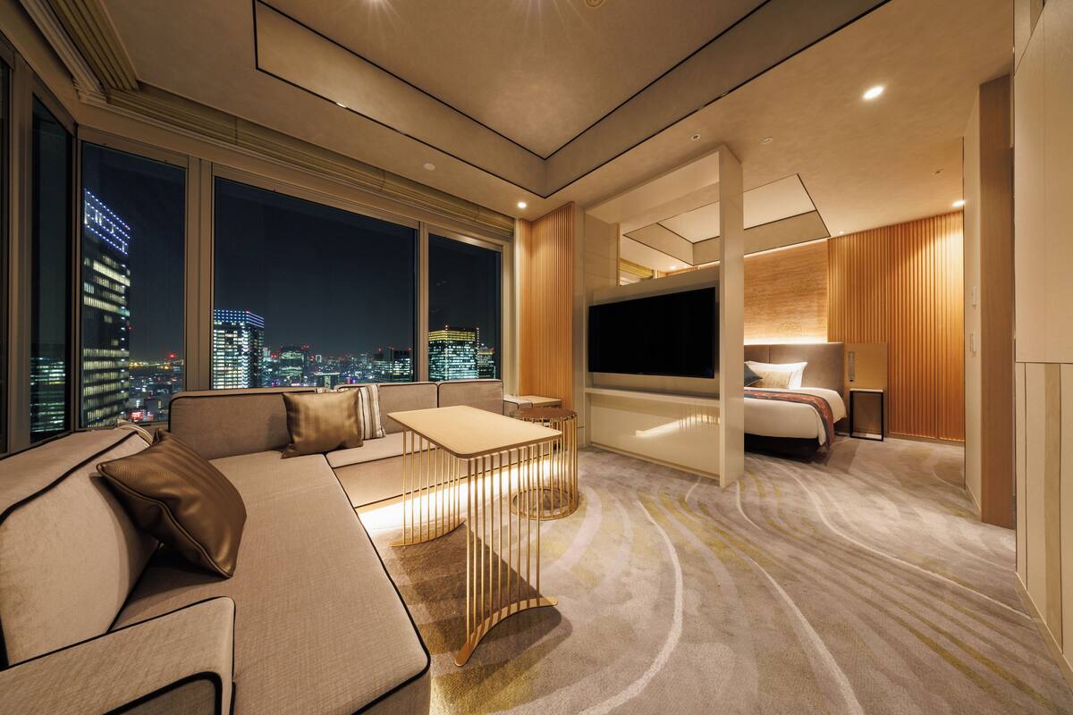 A modern hotel room with a large bed, a sofa, a TV, and a city view.