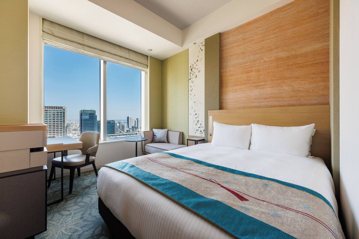 A hotel room with a large bed, a desk, a chair, and a view of the cityscape.