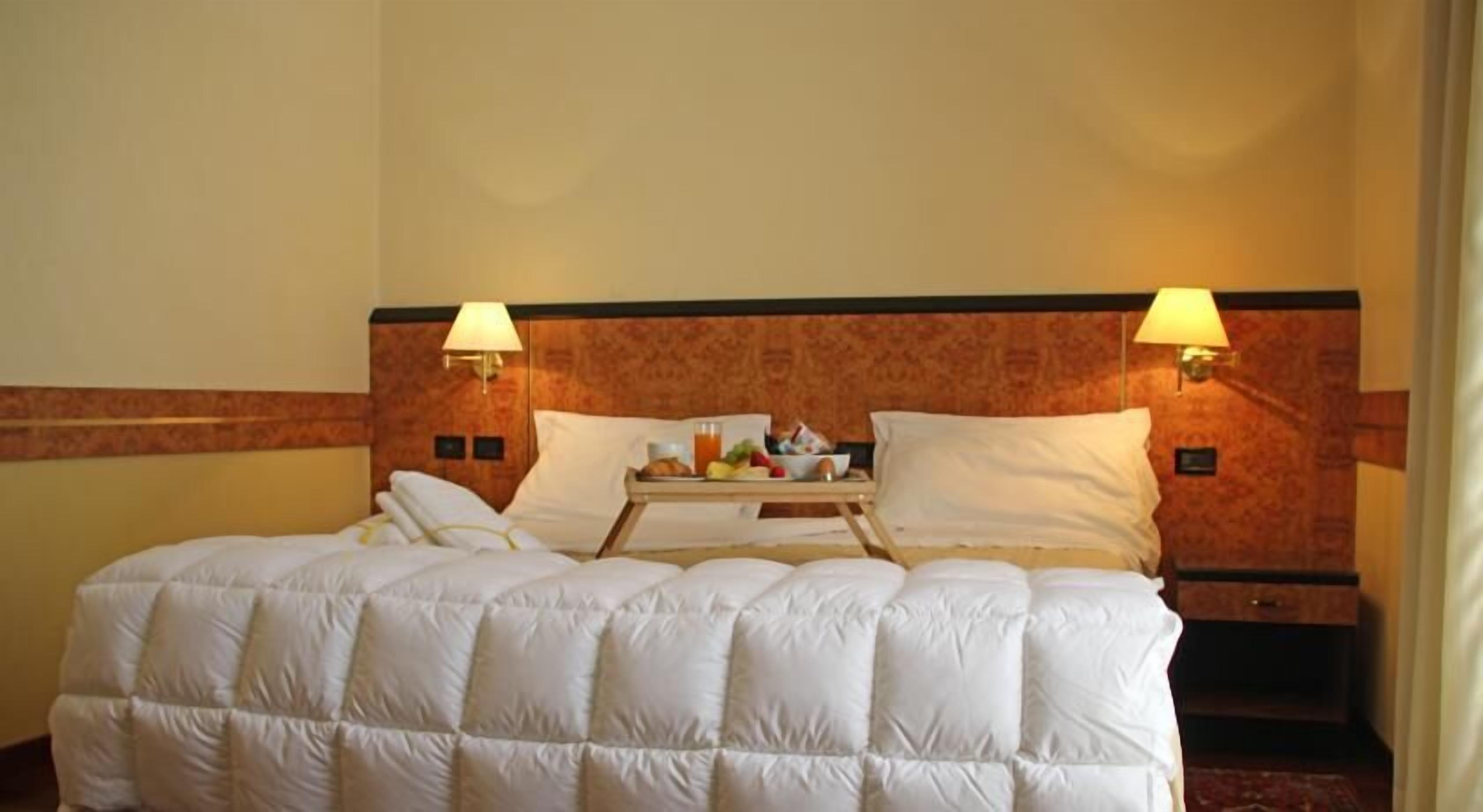 Ein Hotelzimmer mit einem Bett, zwei Nachttischlampen, einem Tablett mit Frühstücksgegenständen und einem Kopfende mit einem gemusterten Design.