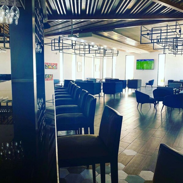 A modern waiting area with rows of chairs, a large screen displaying a sports game, and a bar area with glasses on the left.