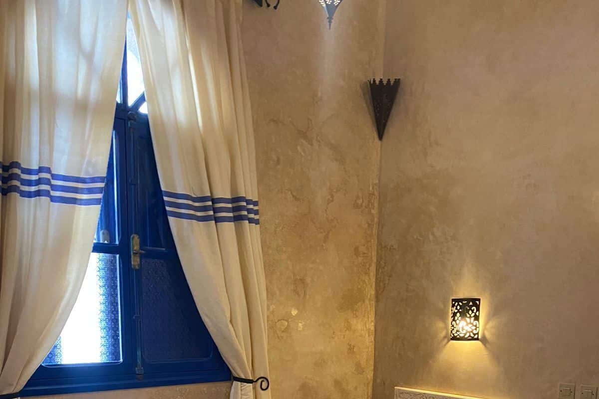 Una cama individual con cabecera, una mesita de noche, una lámpara de pared y una ventana con cortinas.