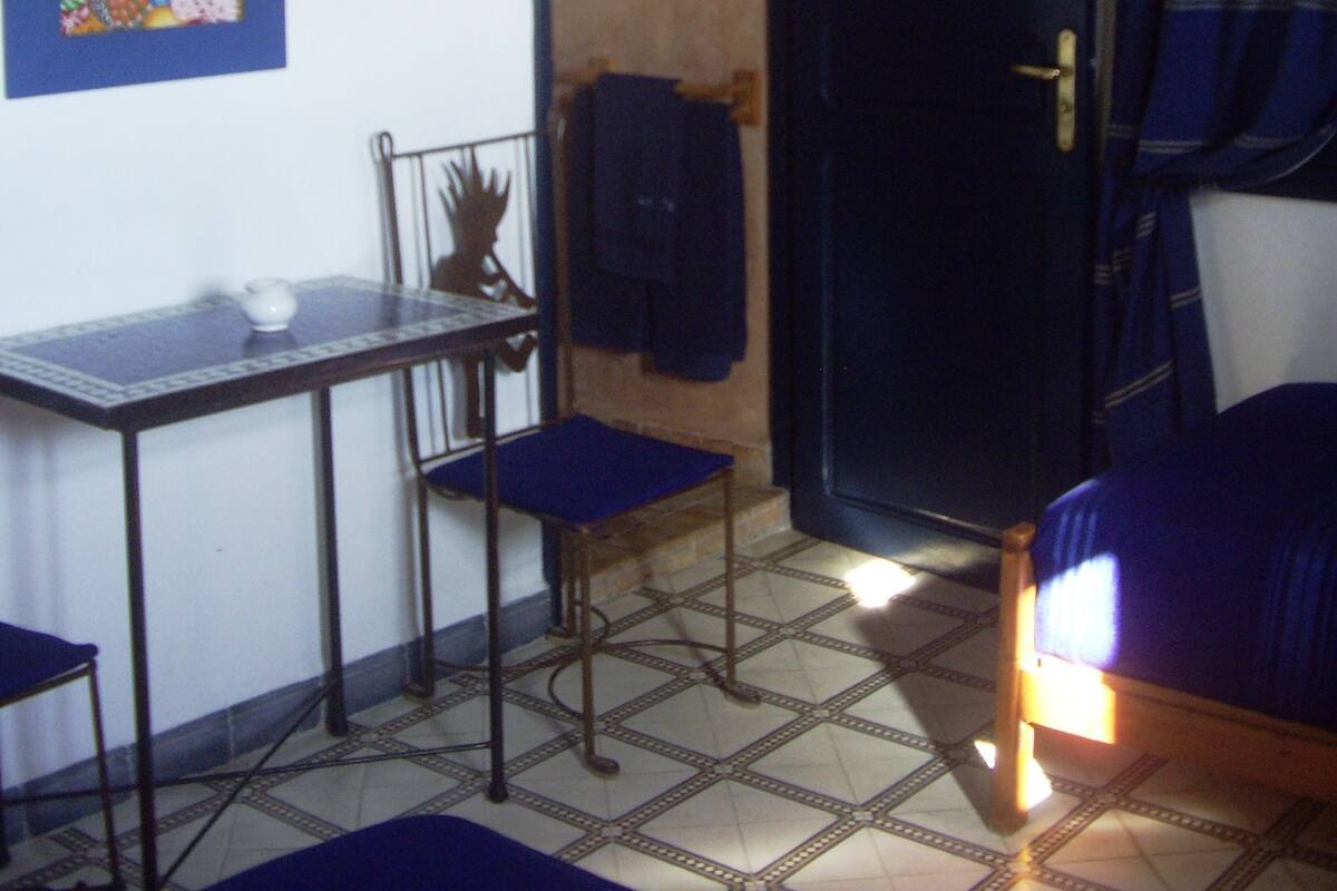 Una habitación con cama, una mesa con silla, una lámpara en la pared, una puerta y un cuadro.