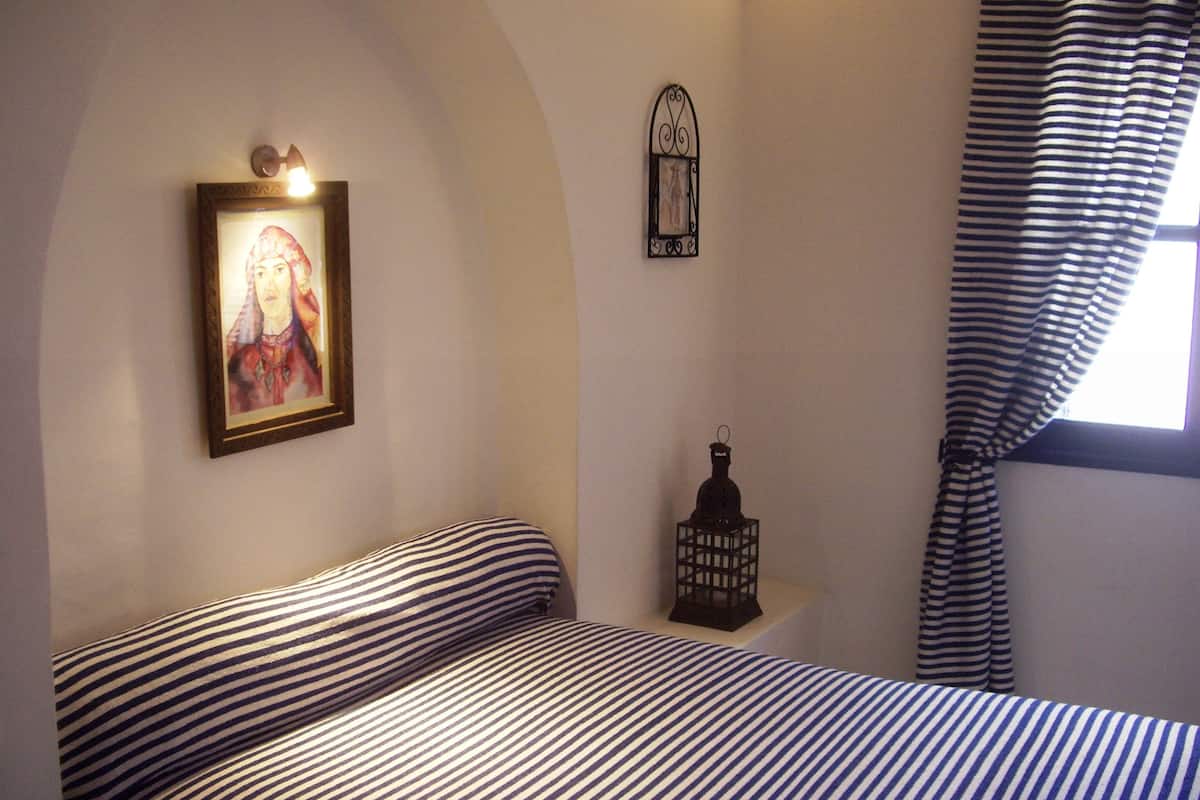 Un dormitorio con cama, un cuadro enmarco, una lámpara de pared, un farol decorativo pequeño y una ventana con cortinas.