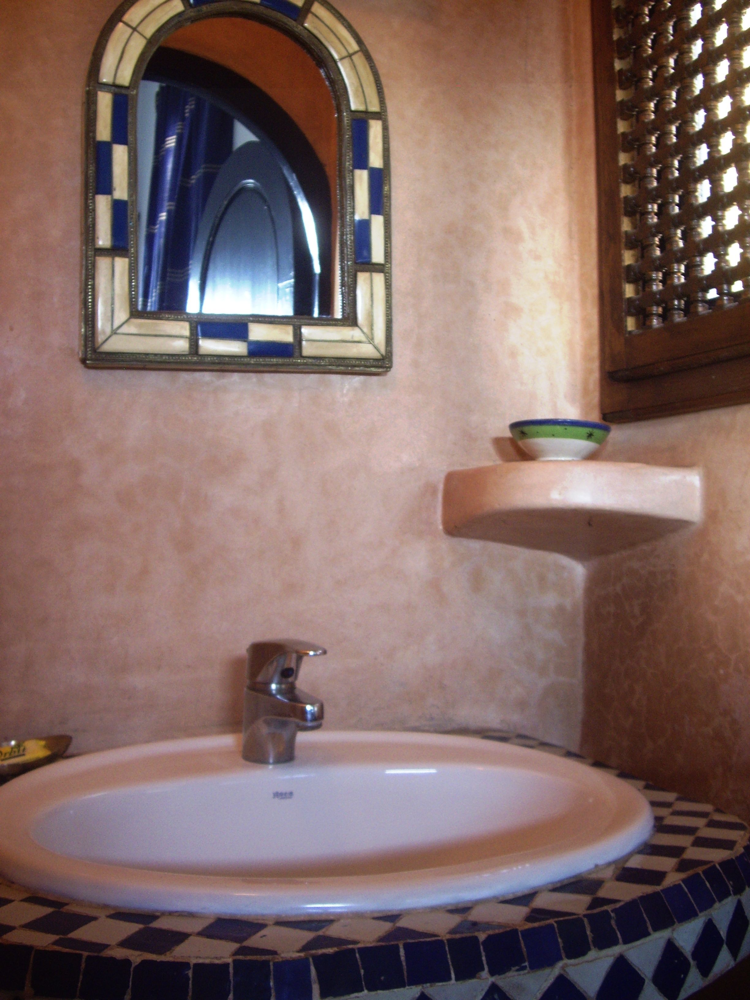 A bathroom with a white sink, a mirror with a mosaic frame, and a shelf with a bowl.
