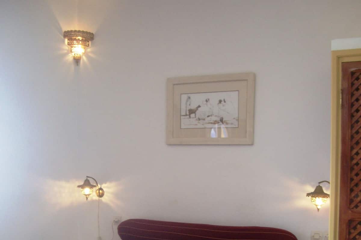 Una cama individual con una colcha estampada, dos luces fijadas a la pared, un cuadro enmarcado y una mesita con estampado de cuadros.