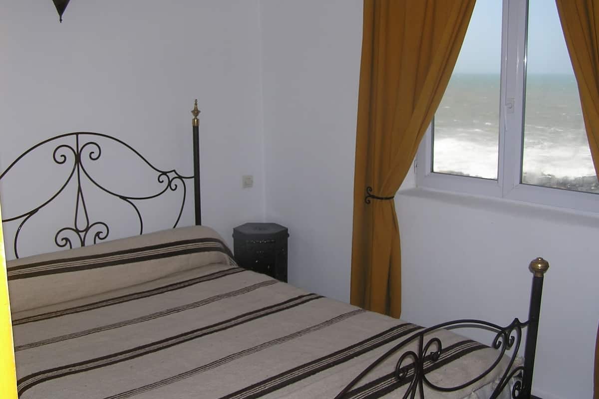 Una cama con colcha a rayas, una mesita de noche, una ventana con cortinas y vista al mar.