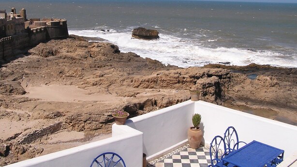 A coastal view with rocky terrain, a white building with blue chairs, and a potted plant.