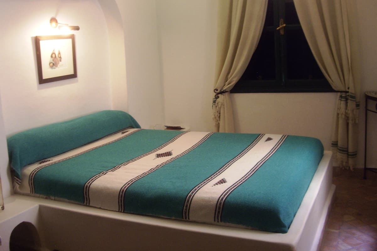 Un dormitorio con una cama, una mesita de noche, un cuadro en la pared y una ventana con cortinas.
