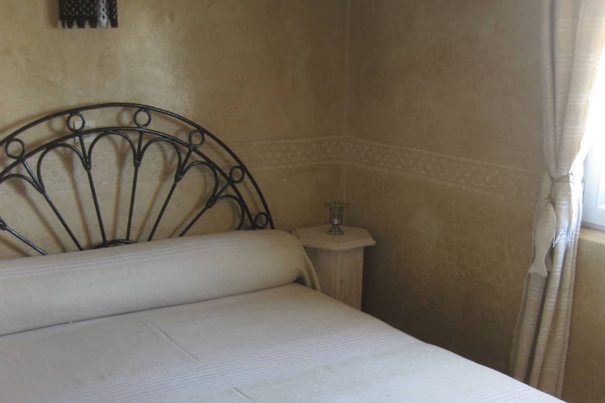 Una cama con cabecera de metal, una mesita de noche con lámpara y un aplique de pared.