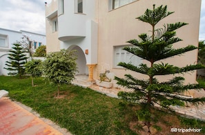 A modern building with a white facade, a small garden with various trees, and a stone pathway leading to the entrance. - Hotel Residence Romane (Hammamet)