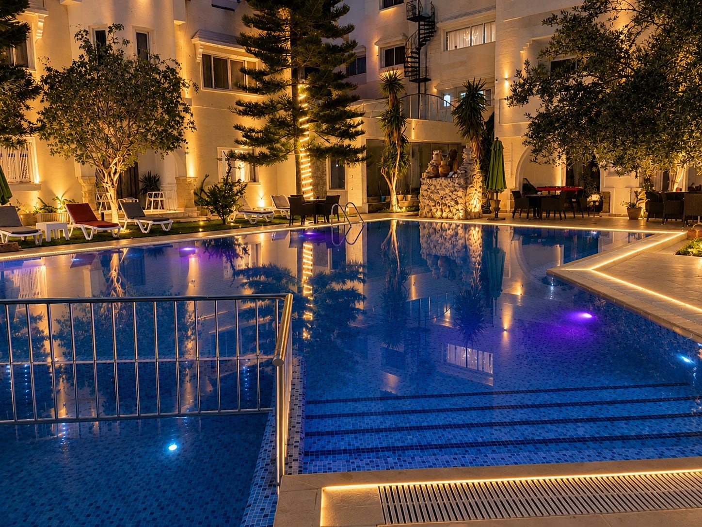 A swimming pool with a tiled edge, surrounded by lounge chairs and potted plants, with a building in the background.
