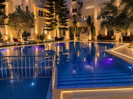 Una piscina con borde de azulejos, rodeada de sillones de descanso y plantas en macetas, con un edificio al fondo.