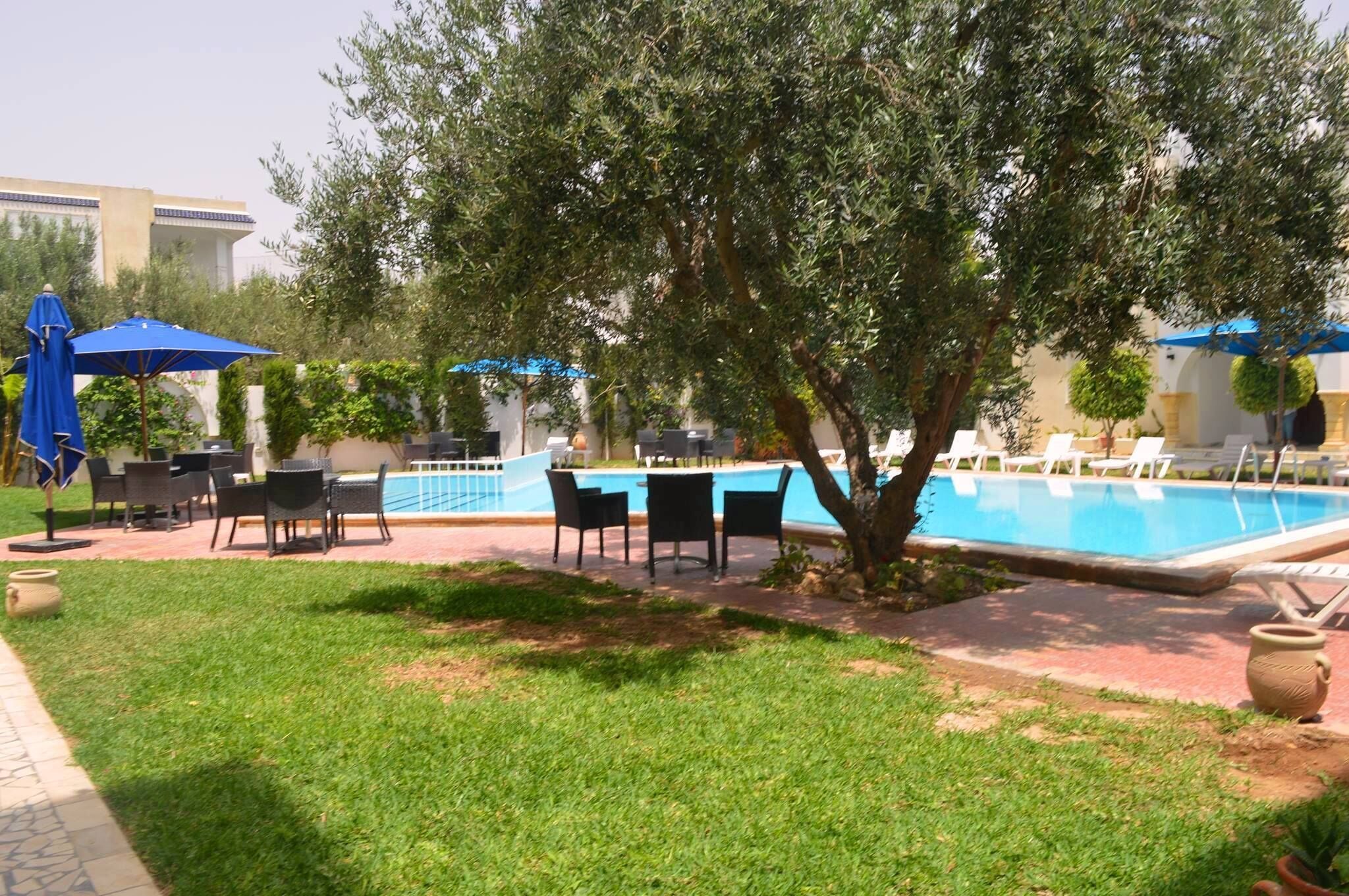 Un espace piscine avec des chaises longues et des parasols, entouré de verdure et avec un bâtiment en arrière-plan.