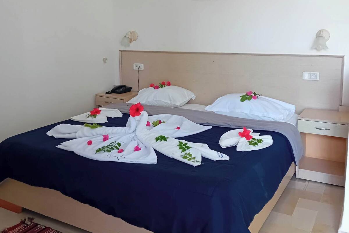 A neatly made bed with white towels and red flowers, a bedside table with a phone, and a small wall decoration.