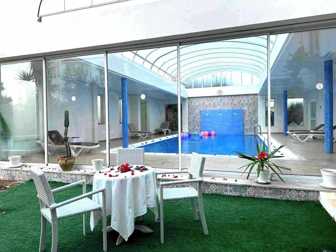Ein von Glas umschlossener Außenbereich mit Pool, weißen Korbmöbeln und einem grünen Rasen.