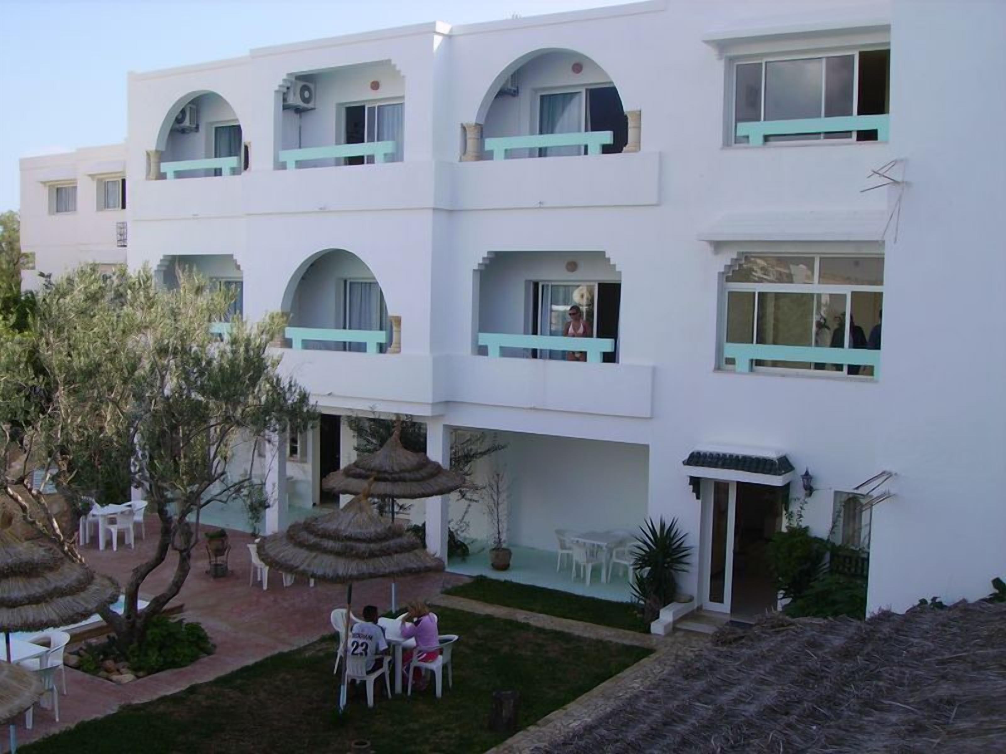 Un bâtiment blanc doté de nombreux balcons, d’un espace extérieur aménagé avec des sièges et d’un petit jardin agrémenté de parasols en chaume.