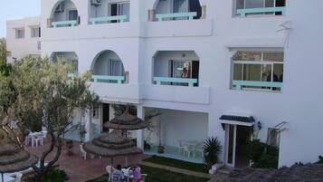 Un edificio blanco con varios balcones, mobiliario exterior y un pequeño jardín con sombrillas de paja.