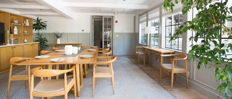 A spacious dining area with a long wooden table and chairs, a kitchen area with wooden cabinets, and large windows providing natural light.