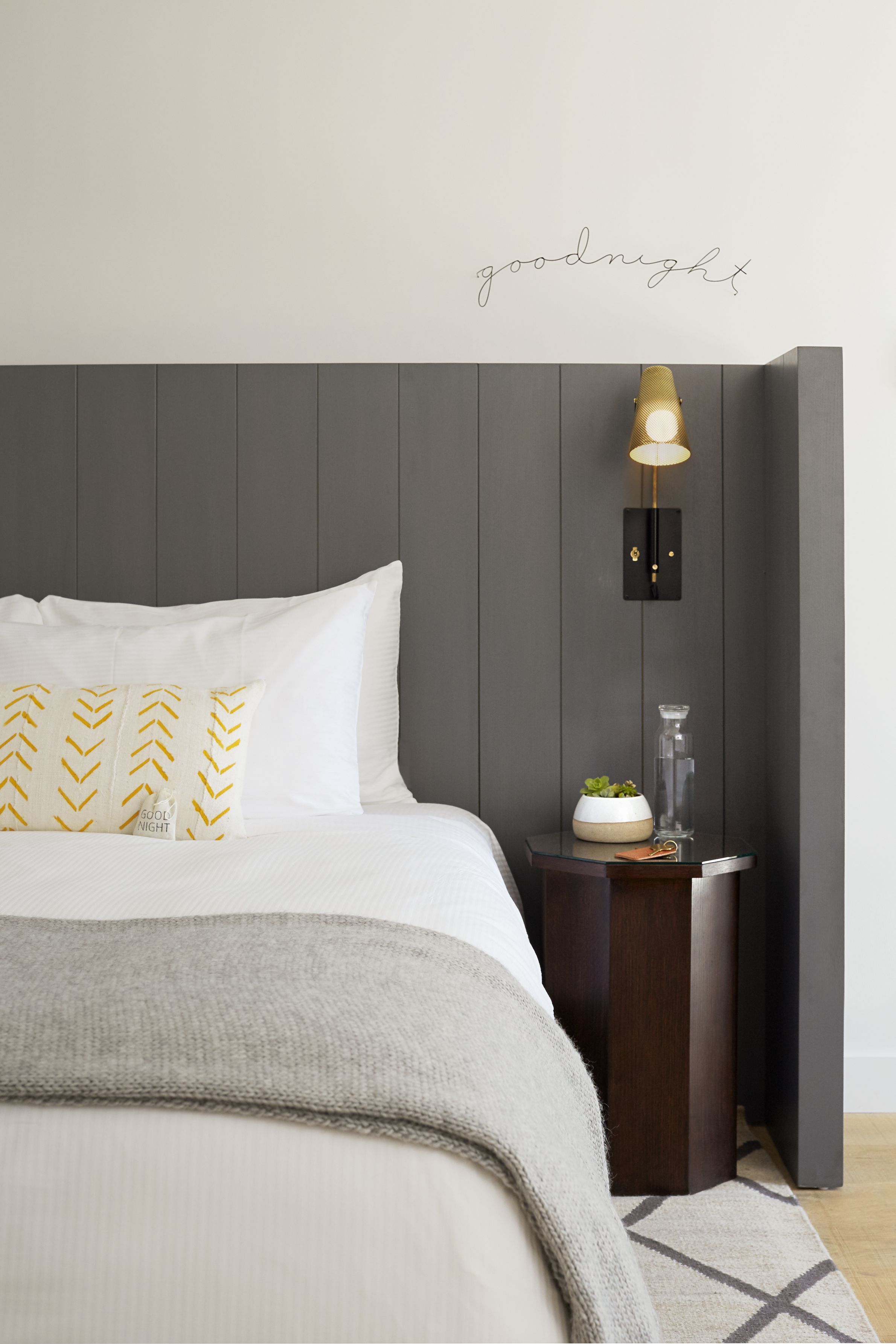 Una cama bien hecha con almohadas blancas y doradas, una mesita de noche con lámpara y una lámpara de pared.
