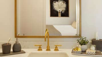 A bathroom with a gold faucet, a mirror, a framed artwork, and toiletries on the counter.