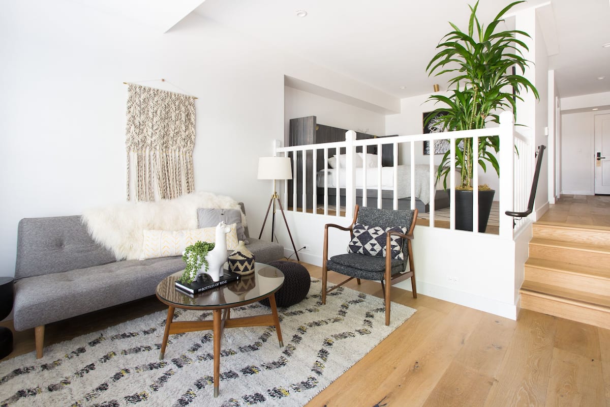 A modern living room with a grey sofa, a round coffee table, and a patterned rug. There is a staircase with a white railing leading to an upper level.