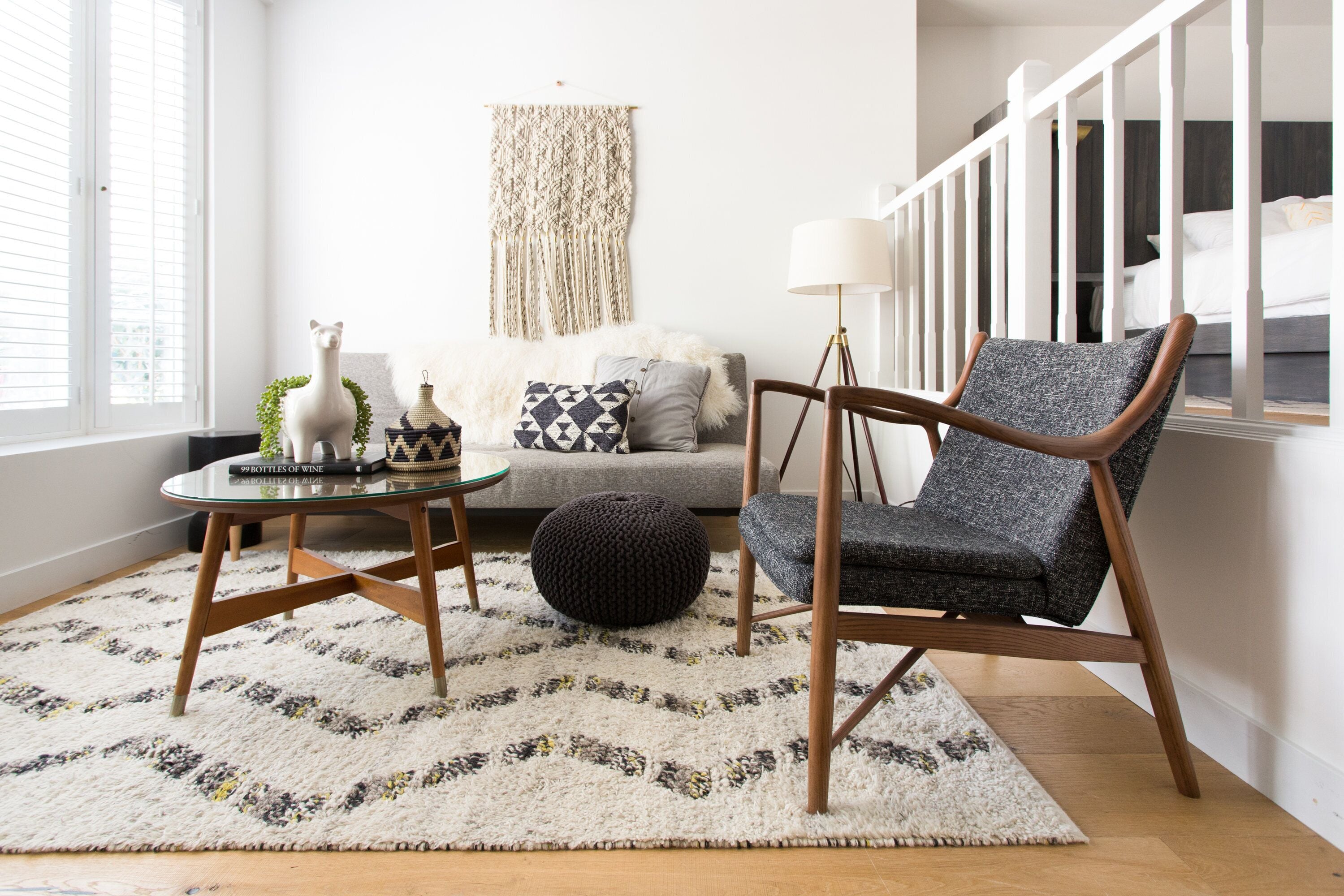 Uma sala de estar moderna com piso de madeira, uma poltrona estofada cinza, um banquinho redondo, uma mesa de centro de vidro e um painel de parede em macramé.